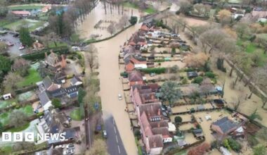 An aerial view of the street, which is partly under a wide streak of brown water. To the left is housing and a church, surrounded by green land and trees, while on the right is more housing and a stretch of farmland.