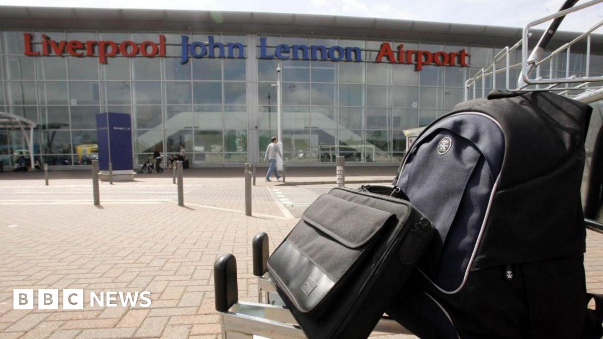 A laptop bag and a backpack sit on a silver trolley in front of Liverpool John Lennon Airport's glass-fronted departures terminal.
