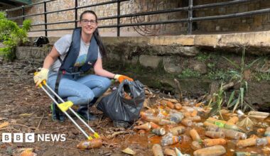A woman wearing glasses, a grey T-shirt and blue jeans, with a life jacket over the top, crouches with a litter picker, next to piles of dirty plastic bottles.