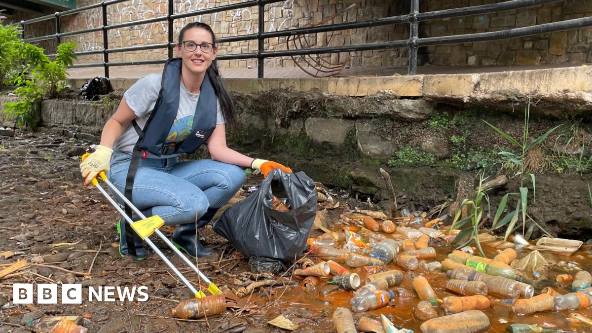 A woman wearing glasses, a grey T-shirt and blue jeans, with a life jacket over the top, crouches with a litter picker, next to piles of dirty plastic bottles.