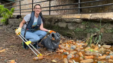 Sheffield City Council A woman wearing glasses, a grey T-shirt and blue jeans, with a life jacket over the top, crouches with a litter picker, next to piles of dirty plastic bottles.