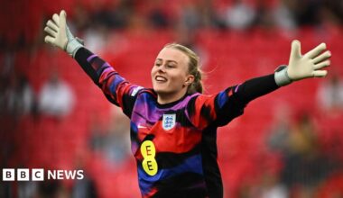 Hannah Hampton in a colourful red, purple, blue and black goalie's jersey, with her hands stretched out wide and a smile on her face. Her blonde hair is tied bak and she is wearing white goalie gloves. Stands are blurred behind her.