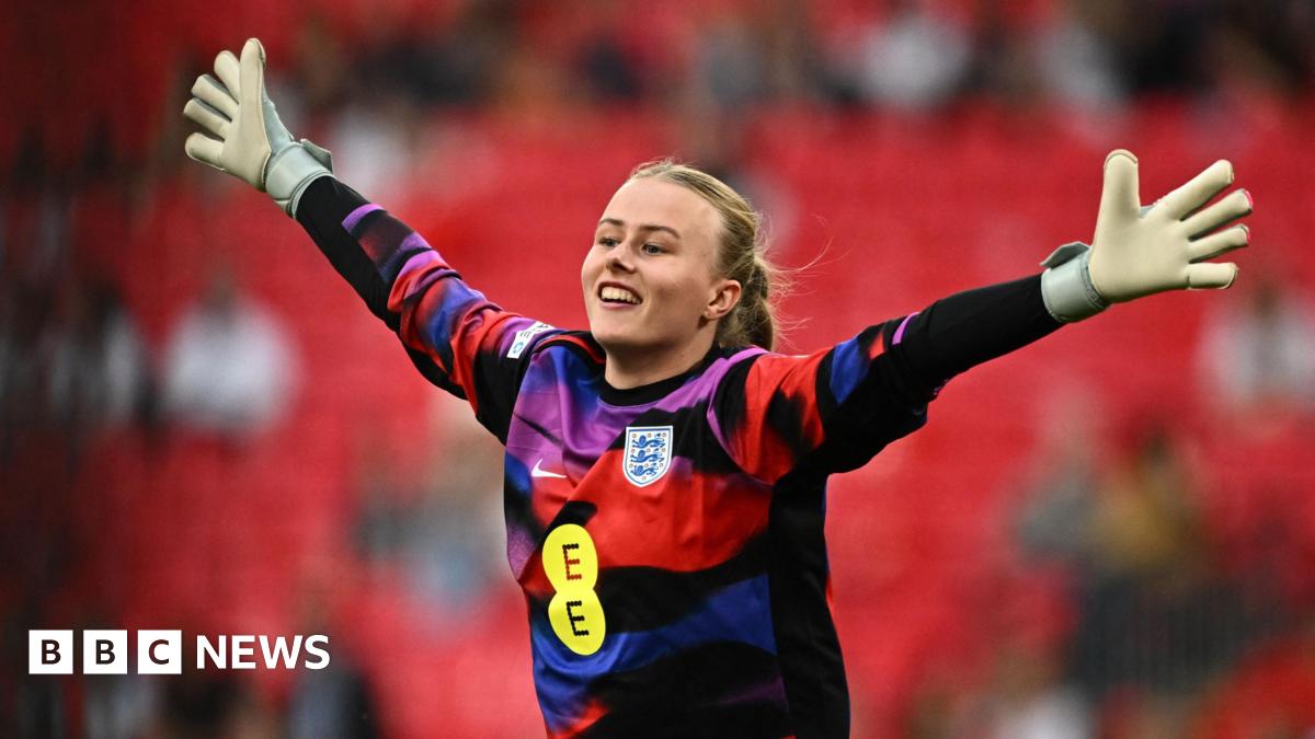 Hannah Hampton in a colourful red, purple, blue and black goalie's jersey, with her hands stretched out wide and a smile on her face. Her blonde hair is tied bak and she is wearing white goalie gloves. Stands are blurred behind her.