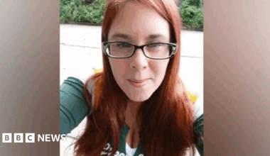 A woman with red hair, dark rimmed glasses and a green and white top is looking towards a camera. There is greenery in the background.