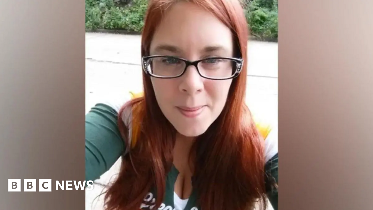 A woman with red hair, dark rimmed glasses and a green and white top is looking towards a camera. There is greenery in the background.