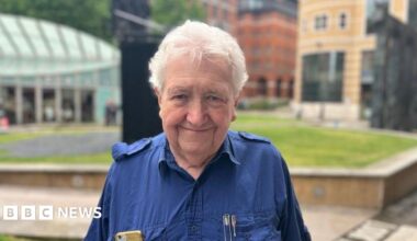 A man with white hair is smiling at the camera. He is wearing a navy blue shirt with his gold mobile phone in one breast pocket and some pens in another.