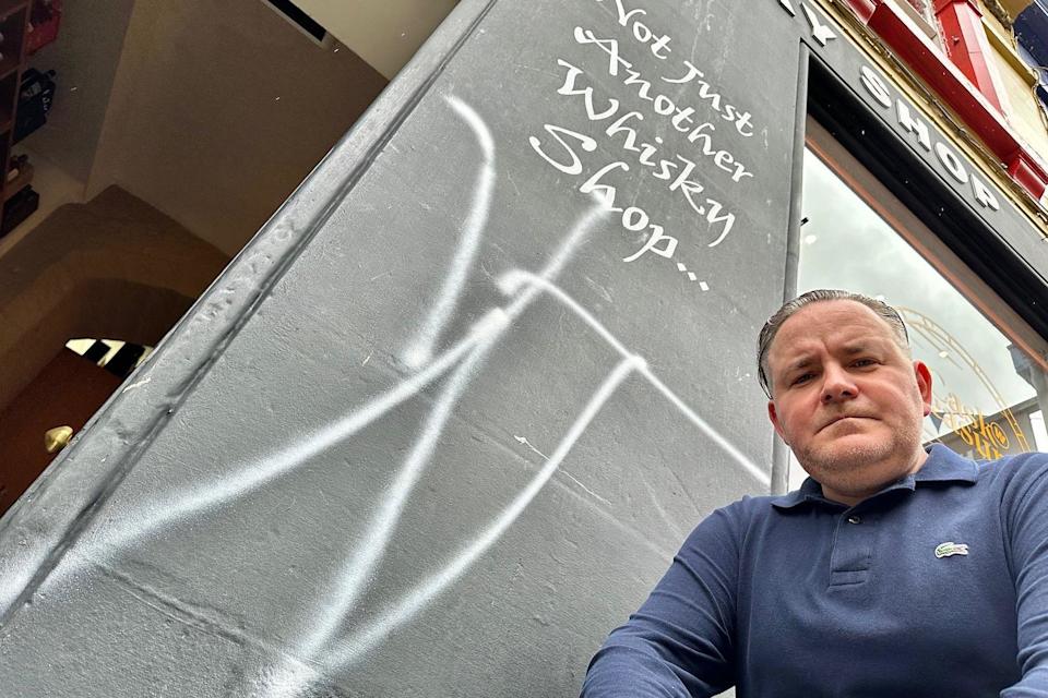 Andrew is looking down at the camera. He is wearing a blue top. There is white graffiti beside him on a shop.
