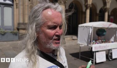 A man with long grey hair holds up a green vape. Behind him is a vape sculpture