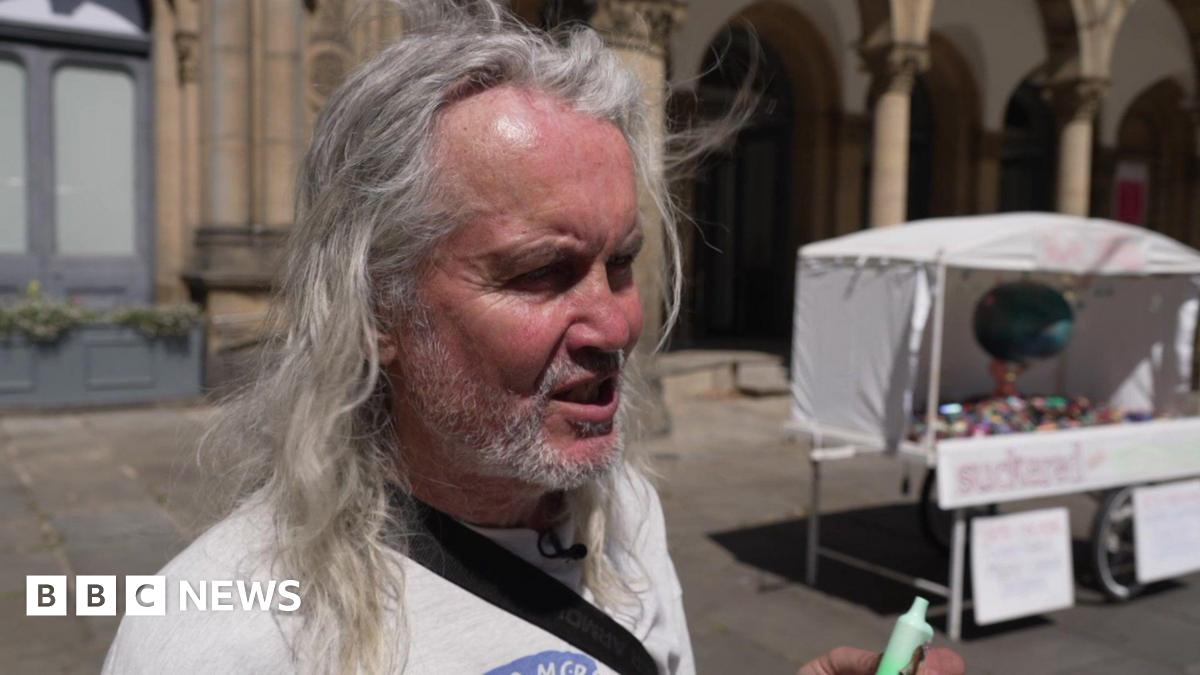 A man with long grey hair holds up a green vape. Behind him is a vape sculpture