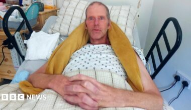 Tim Hull lies in bed with a yellow blanket round his neck and shoulders. He has blue eyes and short hair with his hands folded across his lap