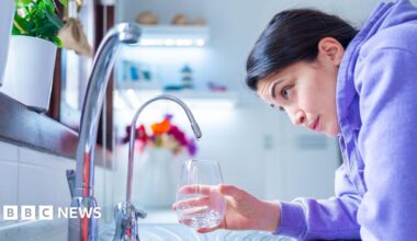 Young woman in a purple hoodie leans over a kitchen sink, holding a glass under a mixer tap. She looks slightly concerned at the lack of water flow from the tap. There is another tap in the foreground.