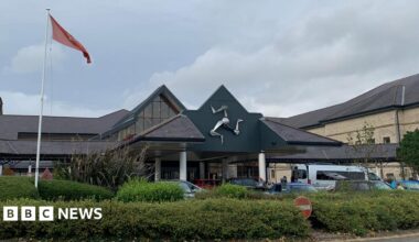 The entrance to Noble's Hospital, with the silver triskelion emblem on the front of the building and a Manx flag in front to the left above a green area of bushes and shrubbery.
