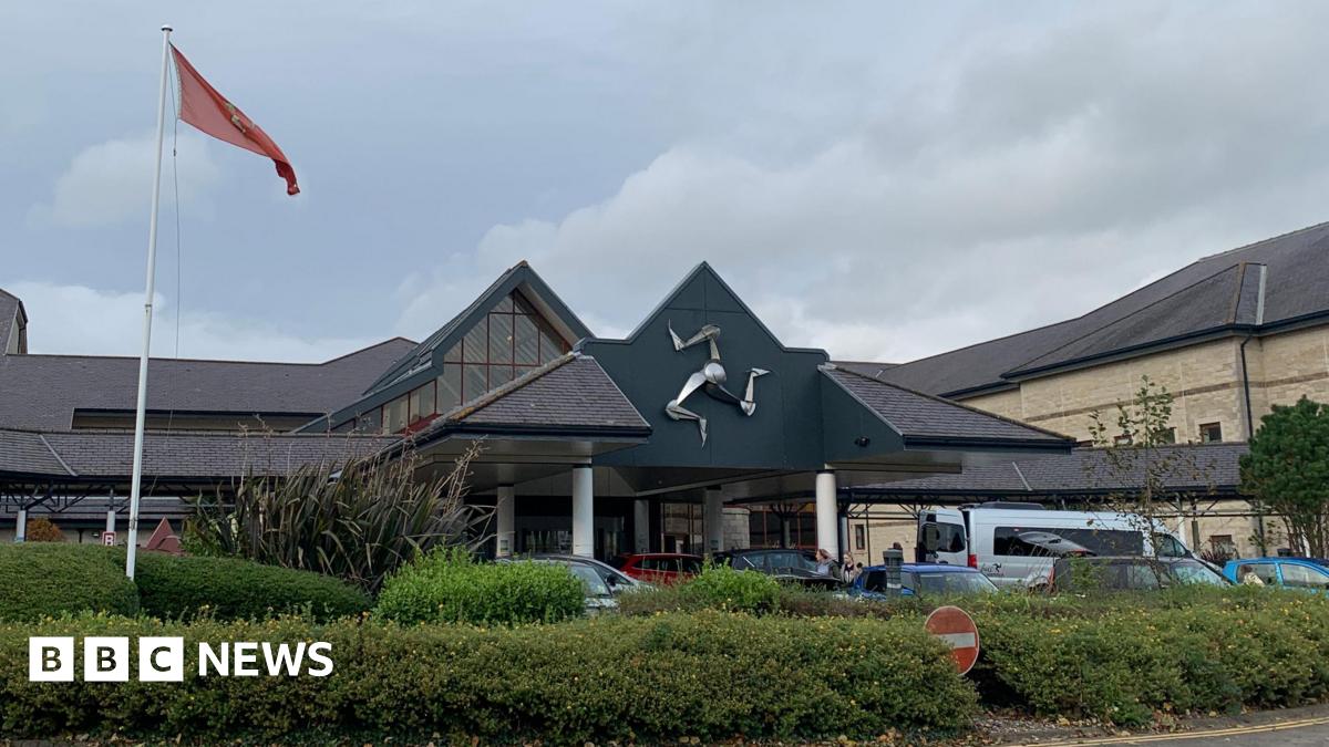 The entrance to Noble's Hospital, with the silver triskelion emblem on the front of the building and a Manx flag in front to the left above a green area of bushes and shrubbery.