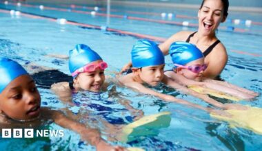 Four young children - two girls and two boys - swimming in a pool with a female instructor. All four children are wearing blue swimming caps and the girls have pink goggles on. They are all holding yellow and green floats in front of them.