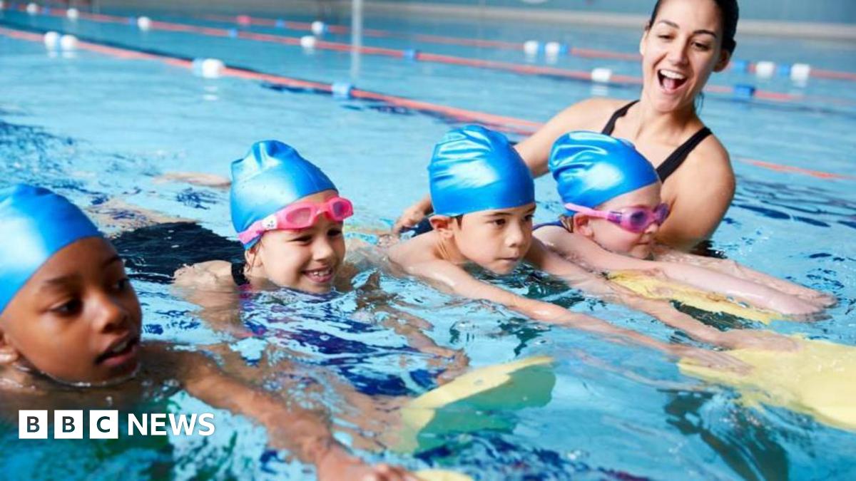 Four young children - two girls and two boys - swimming in a pool with a female instructor. All four children are wearing blue swimming caps and the girls have pink goggles on. They are all holding yellow and green floats in front of them.