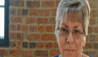Geraldine stares at the camera. She wears a dark blue top and a green lanyard. Behind her is a brick wall with windows either side.