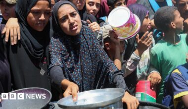 Two women, who are wearing black and have pained looks on their faces, stand at the front of a crowd and hold out pans at a charity kitchen in Nuseirat, Gaza