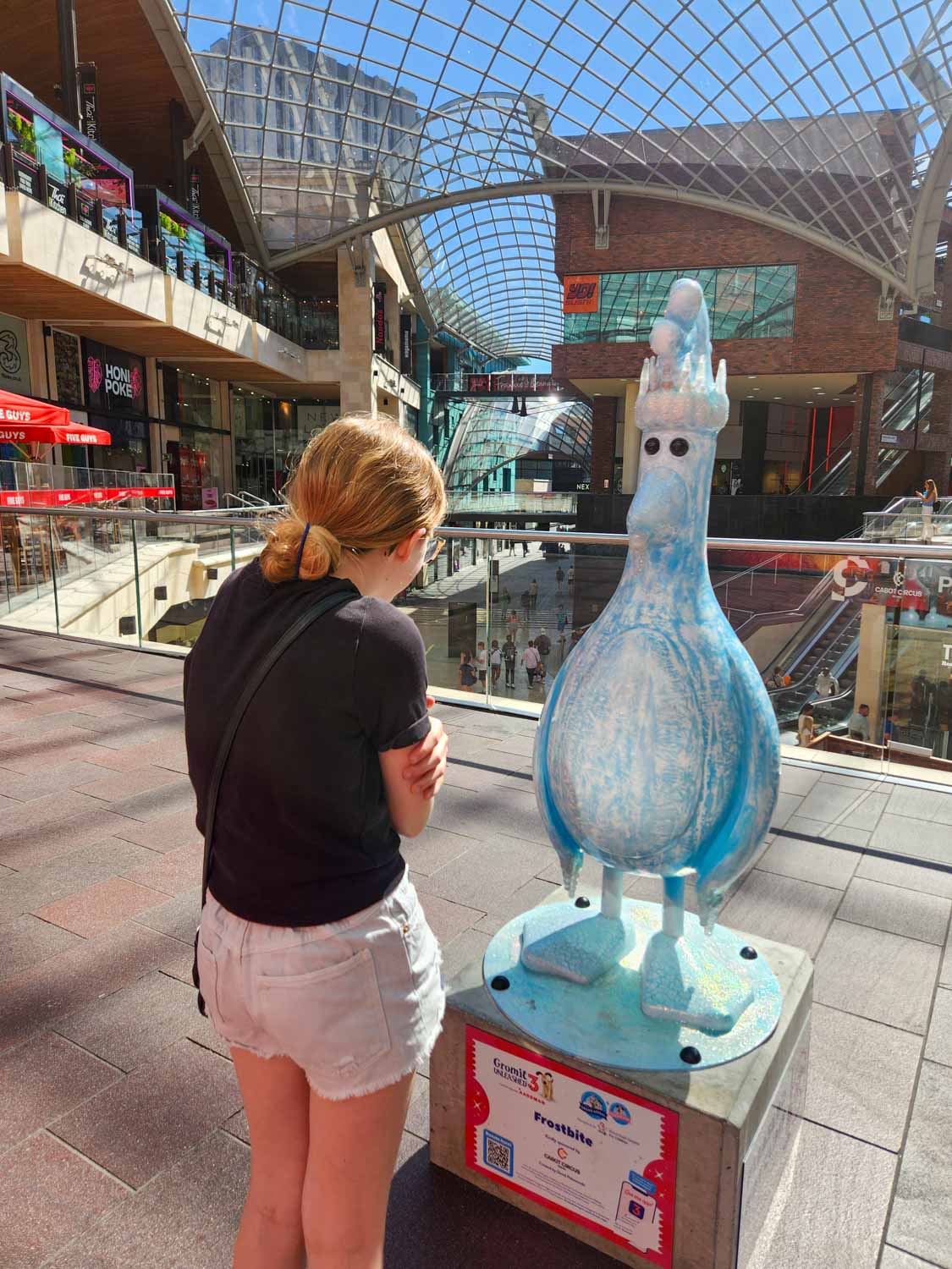 My daughter pretends to shiver next to a statue of a frozen Feathers McGraw - my Gromit Unleashed 3 review of a visit to Bristol with kids