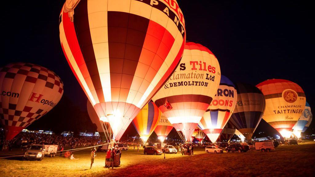 A number of hot air balloons illuminated at night time. They are stationary on a field, with crowds of people looking on.