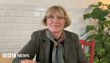 Denise McKay with blonde hair, wearing an olive blazer and a pashmina. She is sitting at a wooden table on a red chair looking at the camera.