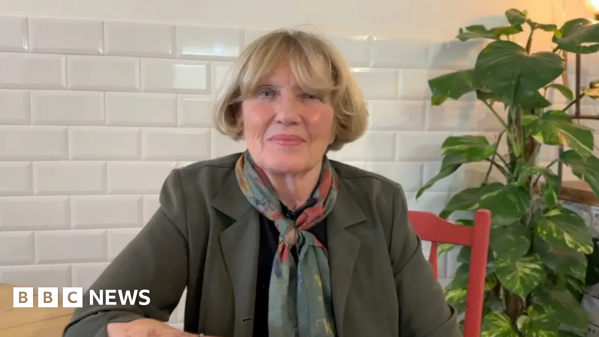 Denise McKay with blonde hair, wearing an olive blazer and a pashmina. She is sitting at a wooden table on a red chair looking at the camera.