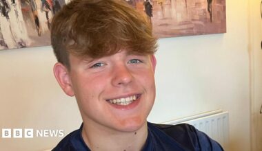 Oscar has brown hair pushed forward, he is smiling. He is wearing a navy blue T shirt with a light blue strip across the chest. He is sat by a table, on which is a certificate, three medals in blue cases and a large silver trophy.