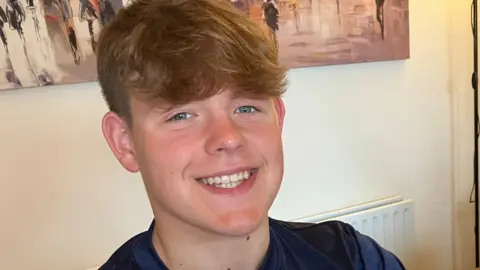 BBC Oscar has brown hair pushed forward, he is smiling. He is wearing a navy blue T shirt with a light blue strip across the chest. He is sat by a table, on which is a certificate, three medals in blue cases and a large silver trophy. 