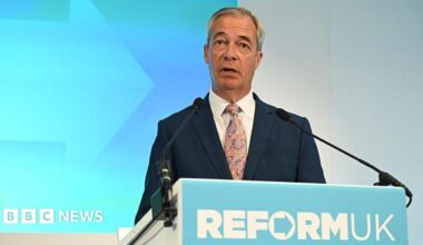 Nigel Farage standing a podium. He is wearing a navy suit and purple and beige tie.  He is standing in front of a light blue and white wall. The podium in front of him reads Reform UK, in the form of the party's logo.