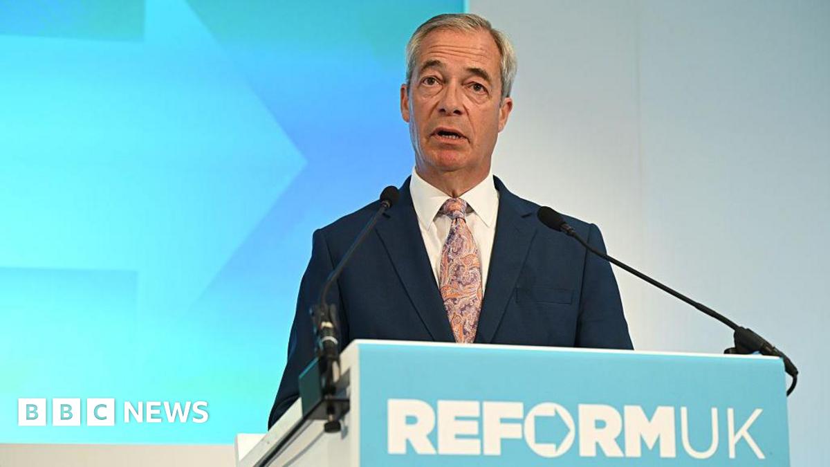 Nigel Farage standing a podium. He is wearing a navy suit and purple and beige tie.  He is standing in front of a light blue and white wall. The podium in front of him reads Reform UK, in the form of the party's logo.