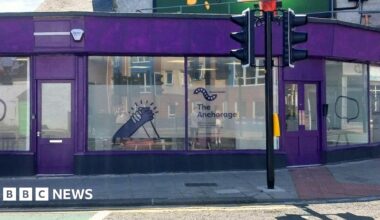 The Anchorage. The drop-in centre is a wide former retail building. It doors and panelling has been painted purple and it has large glass windows all the way around. The middle section of each window has been frosted and decorated with signage for the centre including two hands holding each other.