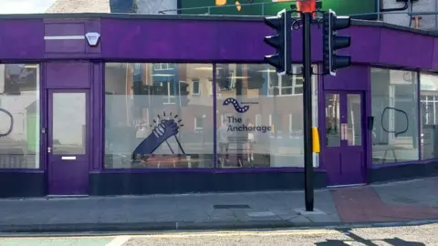 North East and North Cumbria Integrated Care Board The Anchorage. The drop-in centre is a wide former retail building. It doors and panelling has been painted purple and it has large glass windows all the way around. The middle section of each window has been frosted and decorated with signage for the centre including two hands holding each other.