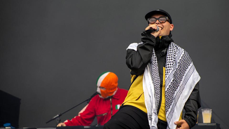 Liam Og O Hannaidh (Mo Chara) from Kneecap performs onstage - he is wearing a keffiyeh, glasses and a baseball cap, while bandmate DJ Provai is behind him on the decks. 
