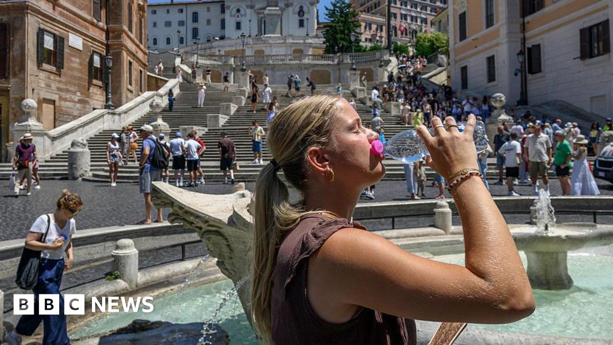 A young woman, her straight light hair pulled into a ponytail, drinks from a plastic water bottle, her eyes closed, in the sunshine. She stands beside a fountain before a staircase, people standing and walking in the background.