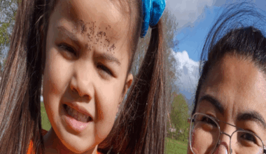 A young girl, with long brown hair in bunches wearing an orange t-shirt, standing in a park next to her smiling mother who is wearing wire framed glasses and has glitter on her face