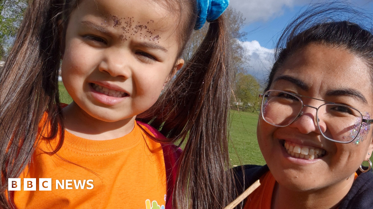 A young girl, with long brown hair in bunches wearing an orange t-shirt, standing in a park next to her smiling mother who is wearing wire framed glasses and has glitter on her face