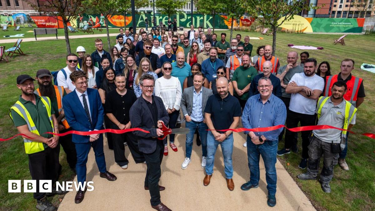A man wearing glasses and a blue blazer holds giant scissors, cutting a red ribbon, in front of large crowd comprising people in work clothing and others dressed casually, in a large green space with a sign reading "Aire Park" in the background.