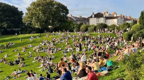 BBC/Steve Mellen A large crowd of people sat on the grass. 