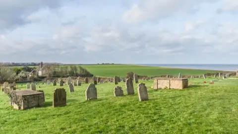 Ian Capper/Geograph A view of a grass graveyard with a number of grave stones. In the distance is the sea, and a house. 