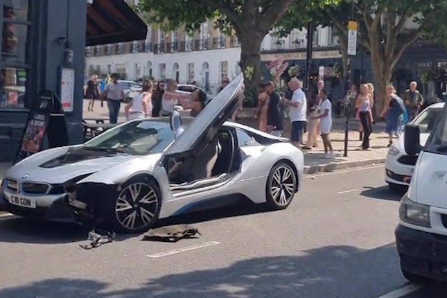 A silver BMW i8 with its doors open after a collision, surrounded by onlookers.