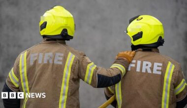 The backs of two firefighters, with one of them having one hand on a shoulder of the other. They are wearing a helmet and mainly brown uniform.
