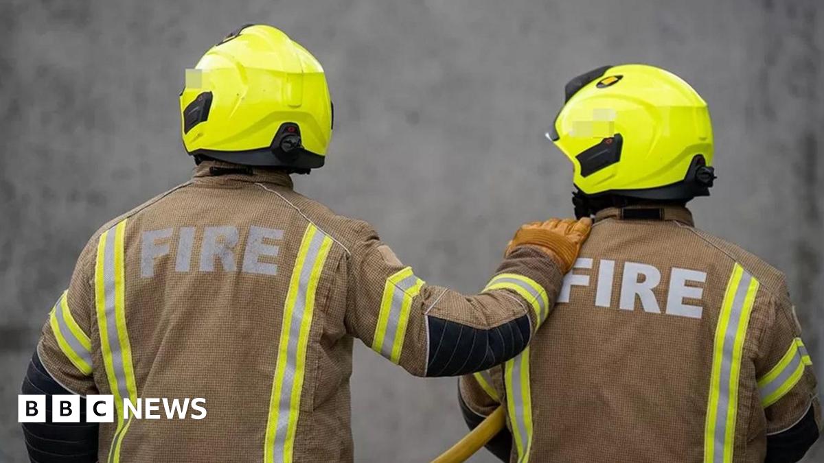 The backs of two firefighters, with one of them having one hand on a shoulder of the other. They are wearing a helmet and mainly brown uniform.