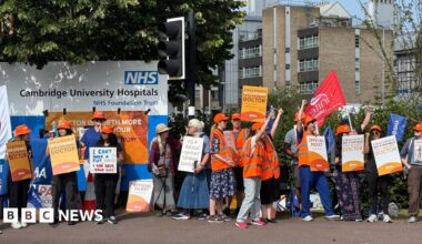 A group of protesters stand with placards and some wearing high-visibility jackets, in front of a sign that reads "NHS: Cambridge University Hospitals". They are holding placards supporting the doctors' strike and some have their hands in the air.