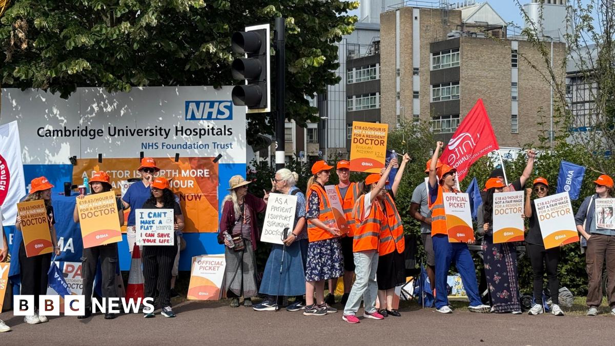 A group of protesters stand with placards and some wearing high-visibility jackets, in front of a sign that reads "NHS: Cambridge University Hospitals". They are holding placards supporting the doctors' strike and some have their hands in the air.