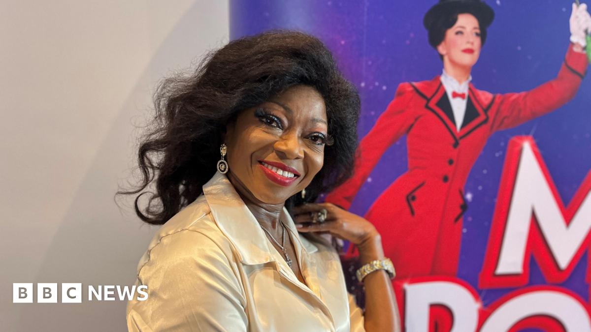Patti Boulaye at the Birmingham Hippodrome. In the head and shoulders shot she is wearing a cream coloured shirt and is smiling.  Behind her is part of a red and purple advertising board for Mary Poppins