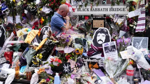 PA Media A man stood surrounded by flowers and notes and a sign saying Black Sabbath Bench