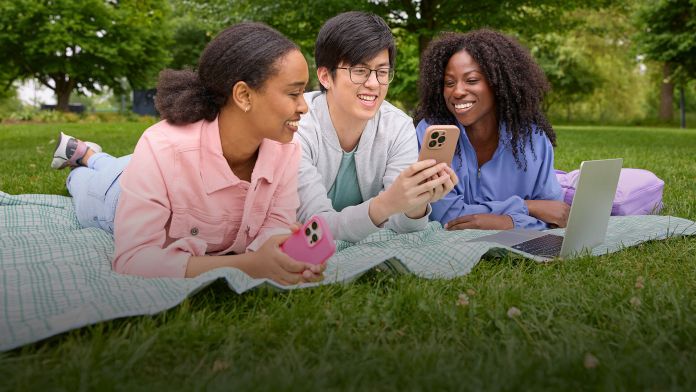 Three young adults sitting on a picnic blanket in a park, engaging with their smartphones and a laptop, surrounded by green grass and trees.