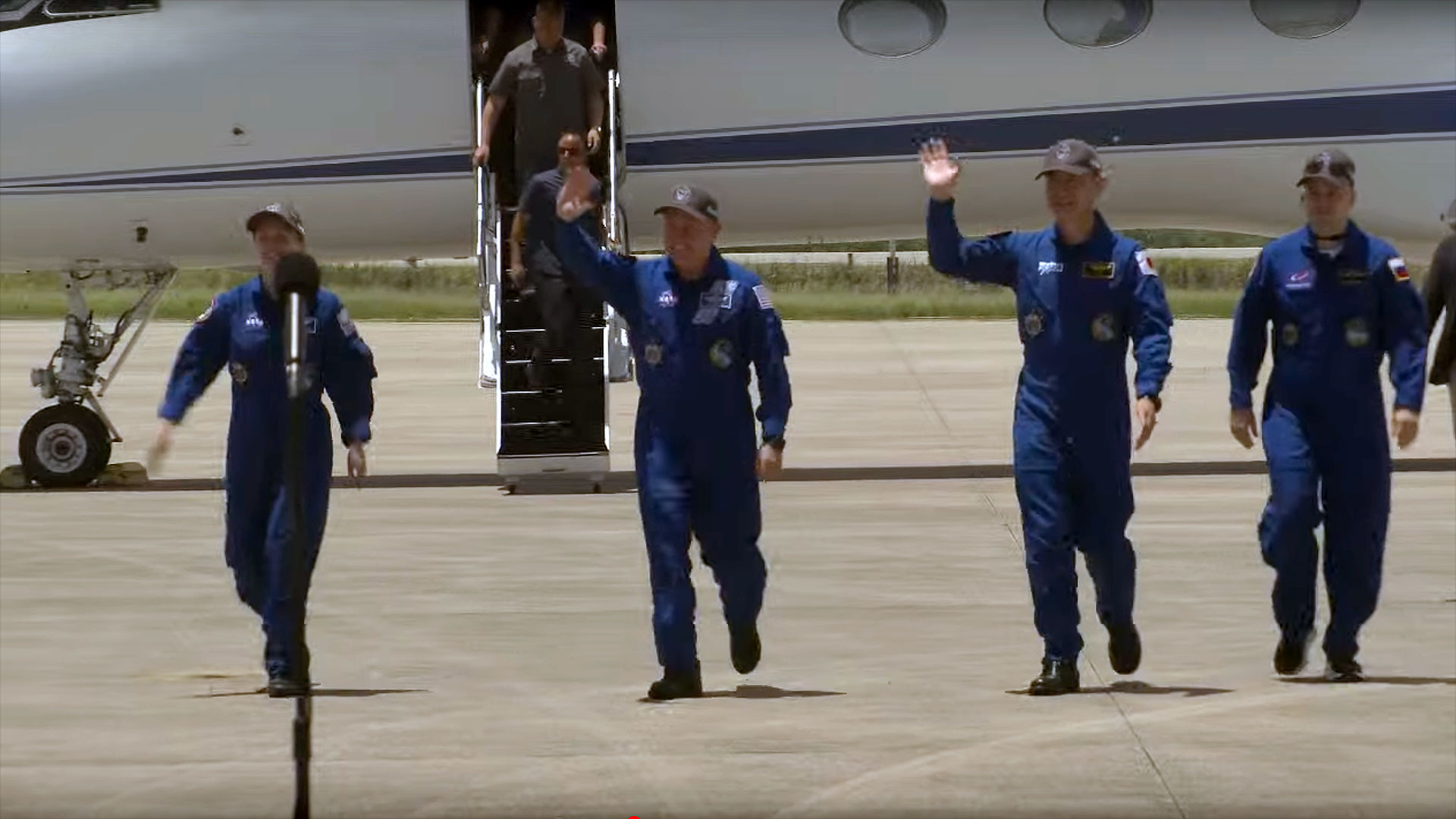 four people in blue flight suits walk on a tarmac toward a microphone stand