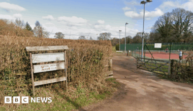 A wooden sign in front of a hedge saying Aston Ingham Cricket Club and Aston Ingham Tennis club. To the right of the picture behind a gate is a red tennis court with a few people standing on the court