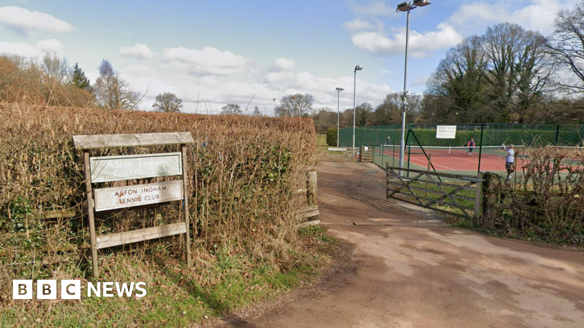 A wooden sign in front of a hedge saying Aston Ingham Cricket Club and Aston Ingham Tennis club. To the right of the picture behind a gate is a red tennis court with a few people standing on the court