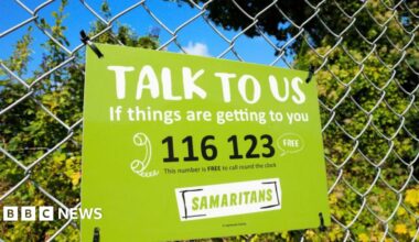Samaritans sign on a railing in front of some bushes. The sign is green with black writing and gives their contact number.